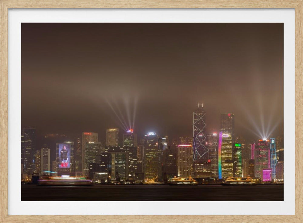 A framed photograph of the Hong Kong skyline at night, viewed from across the water. The skyscrapers are brilliantly lit with yellow, white, and colorful lights, and beams of light shoot up into the foggy, glowing sky. Decor