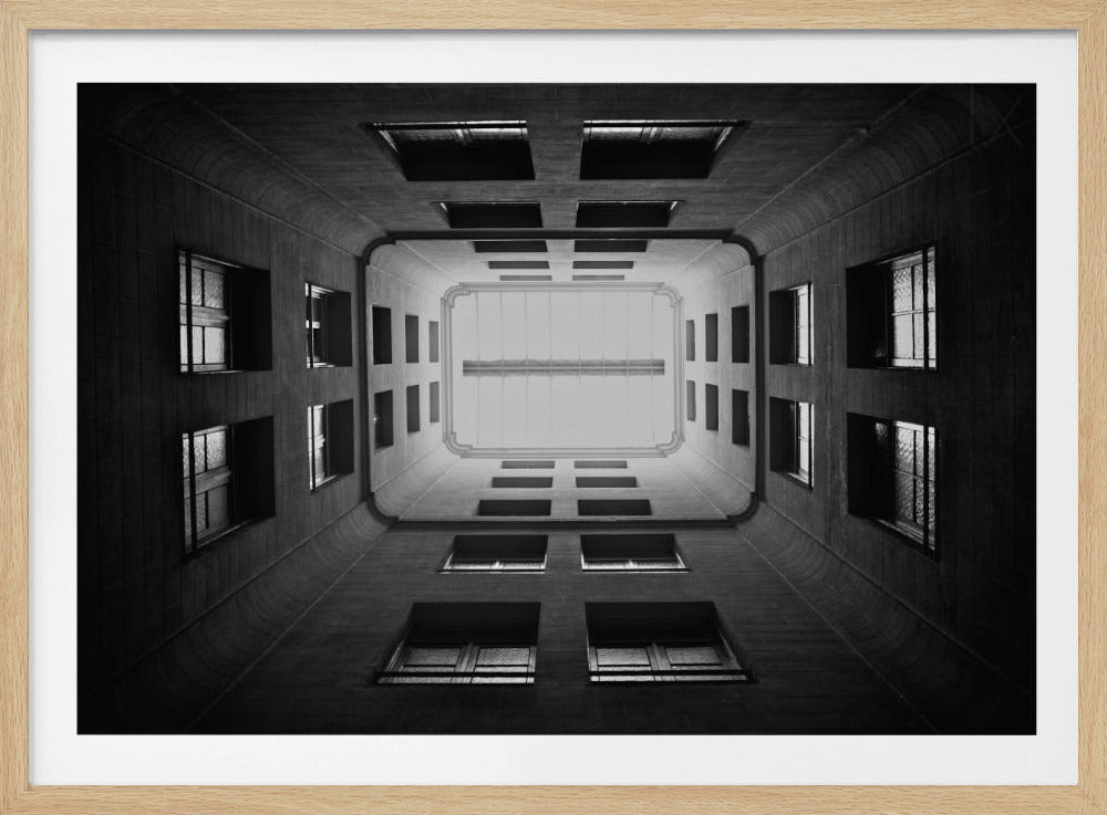 A symmetrical, black and white photograph looking directly up from the base of a building's interior courtyard. The dark walls rise up, lined with rows of windows, creating a powerful sense of perspective that draws the eye to the bright rectangle of the sky or a skylight at the top. The image is enclosed in a simple silver frame. Poster