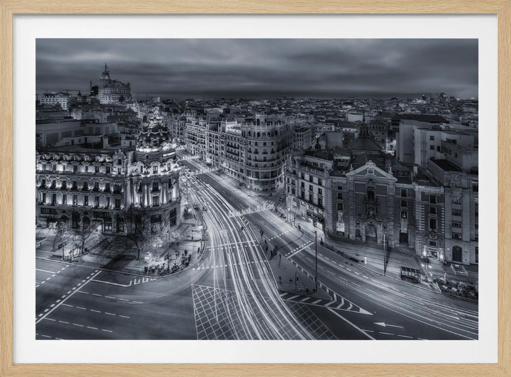A high-angle, black and white long-exposure photograph of a bustling city street at night, possibly Gran Vía in Madrid, enclosed in a silver frame. Light trails from moving vehicles create white streaks on the wide, curving road, which is surrounded by illuminated, ornate historical buildings under a dark, cloudy sky. Artwork