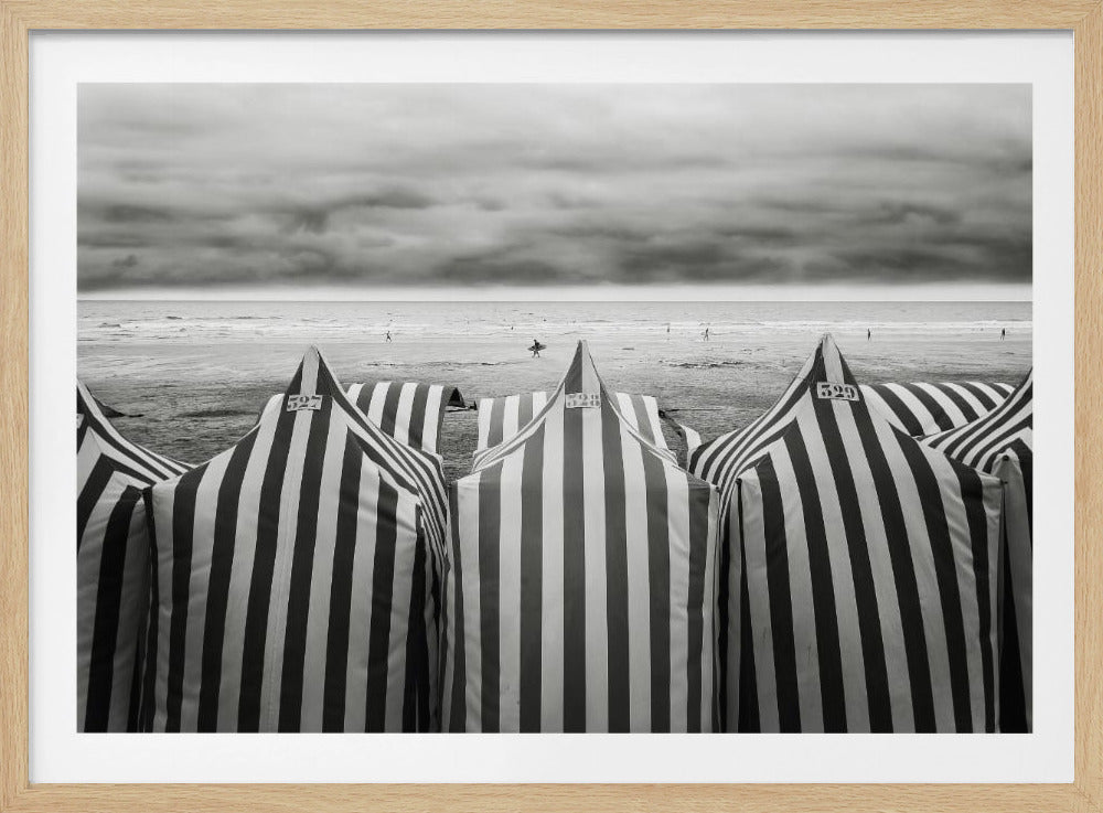 A framed, black and white photograph of striped beach tents on a cloudy day, with the ocean and distant surfers in the background. Print