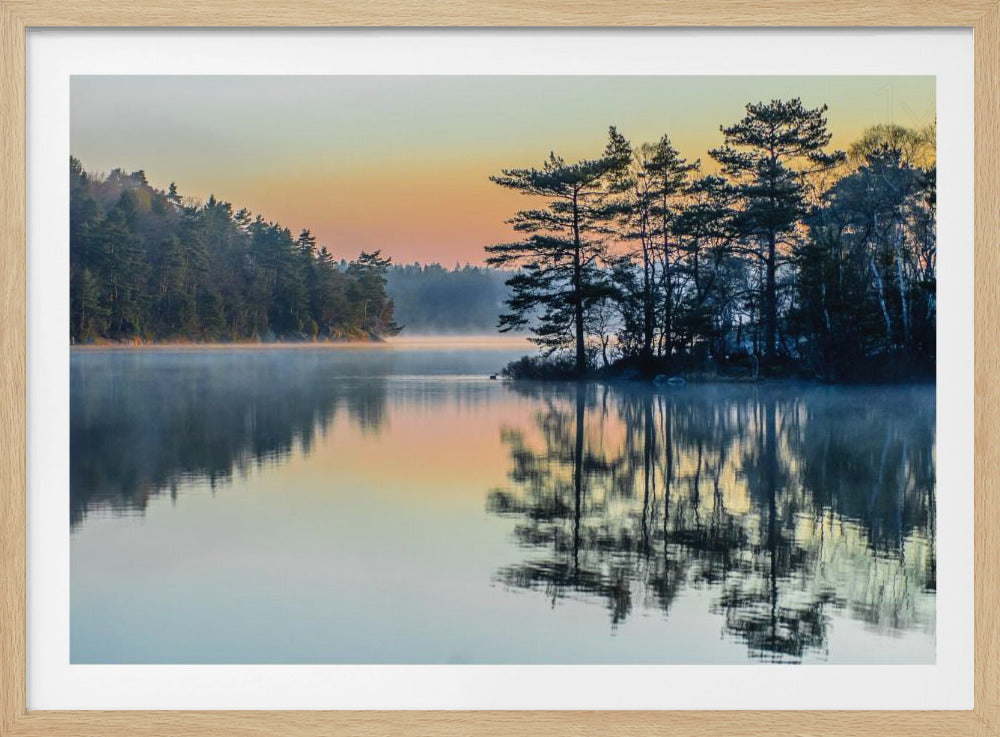 A framed photograph of a serene lake at sunrise. Mist gently rises from the calm water's surface, reflecting a cluster of silhouetted pine trees and the warm orange and yellow glow of the morning sky. A dense forest lines the distant shore. Artwork