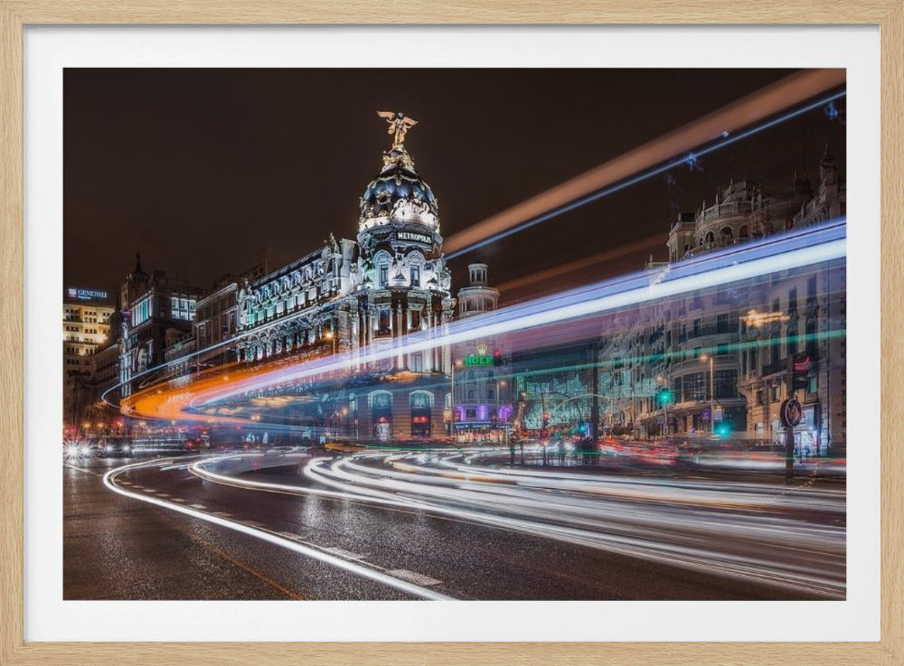 A dynamic long-exposure night photograph of the Metropolis Building in Madrid, with vibrant streaks of white, orange, and blue light from traffic swirling through the city streets. Artwork