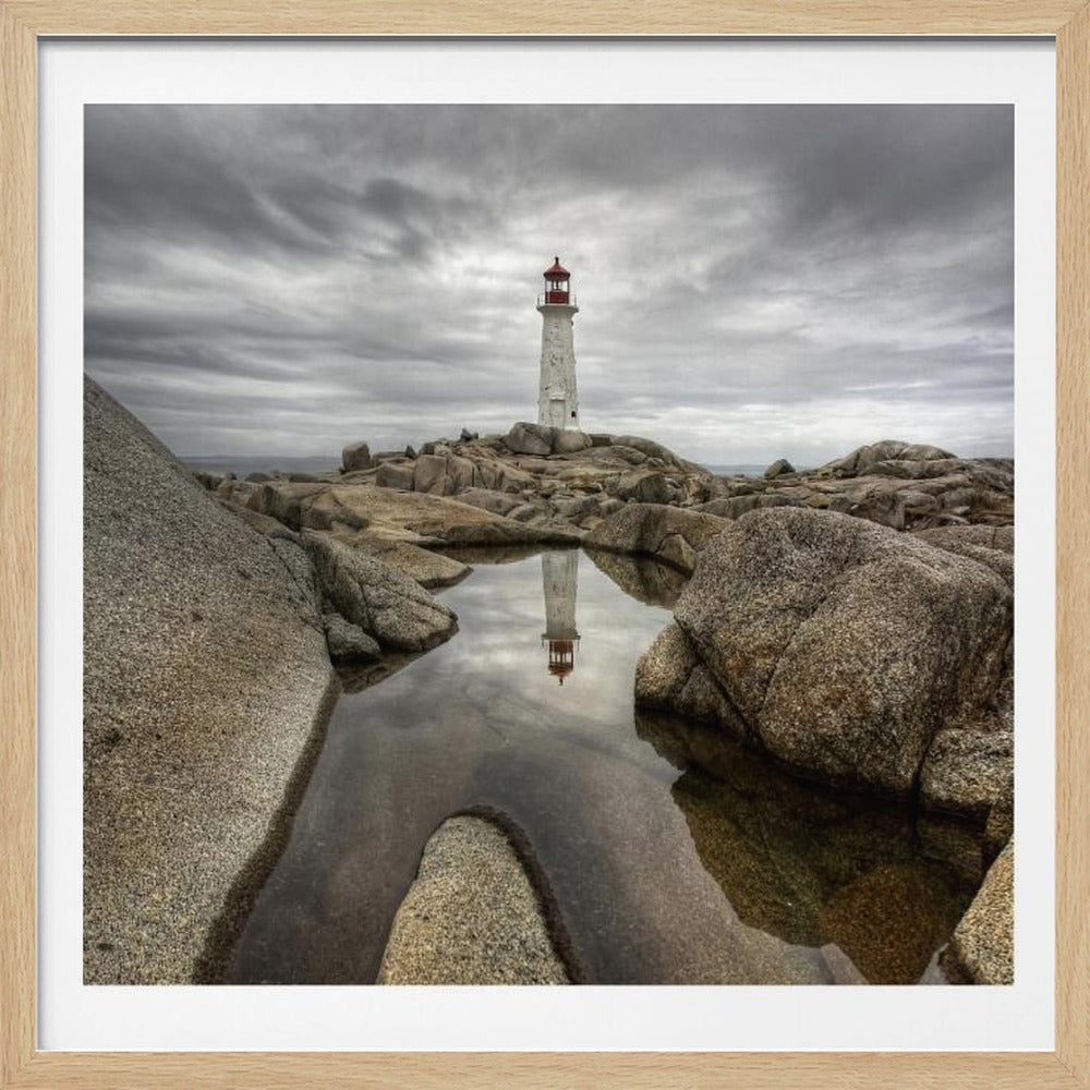 A framed photograph of a white lighthouse with a red top standing on a rocky shore under a dramatic, cloudy sky. In the foreground, a tide pool perfectly reflects the lighthouse. Artwork