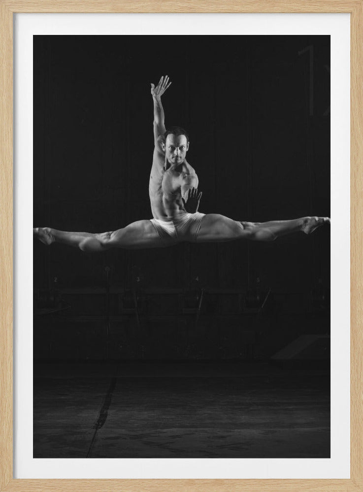 A dramatic black and white photograph of a muscular male ballet dancer frozen in mid-air. He is executing a perfect split jump, his legs extended horizontally. He wears only white briefs, showcasing his defined physique against a dark, moody background. He looks intensely at the camera, with one arm raised elegantly and the other held out in a poised gesture. Artwork