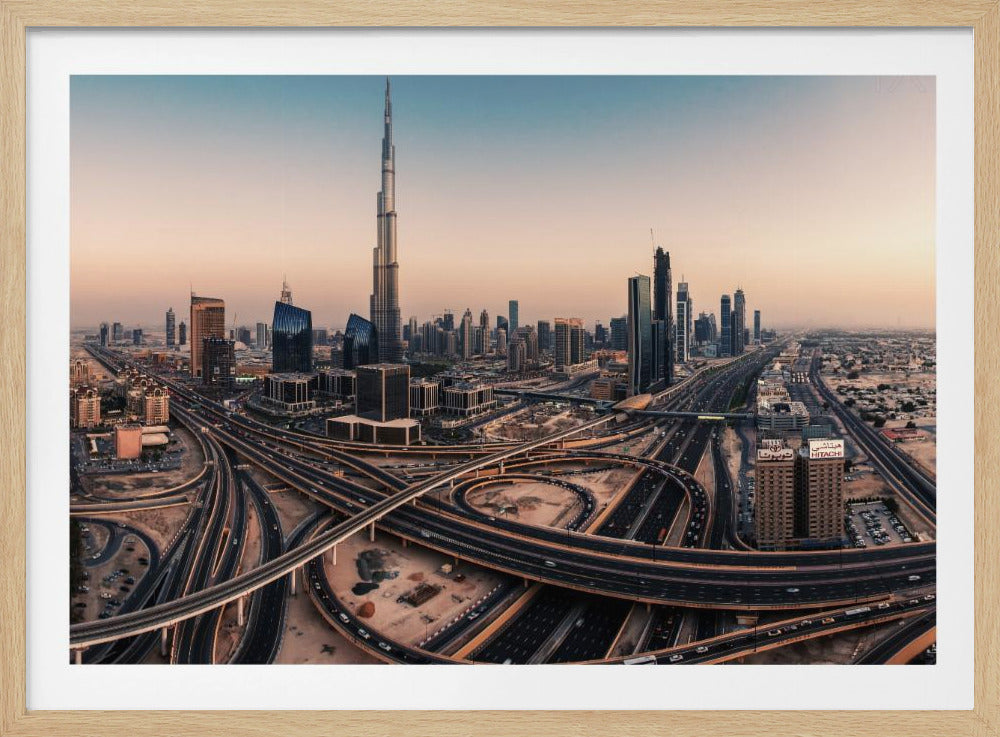 An aerial photograph of the Dubai skyline at sunset, featuring the iconic Burj Khalifa towering over other skyscrapers, with a complex web of intersecting highways and overpasses in the foreground, all under a gradient sky of orange and blue. The photo is displayed within a silver frame. Poster