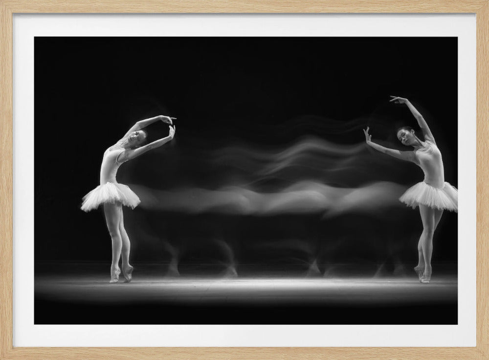 A black and white long-exposure photograph of two ballerinas in white tutus. They are posed elegantly en pointe at opposite ends of a dark stage, connected by a ghostly white motion blur that captures their movement between the two positions. The image has a silver frame. Decor