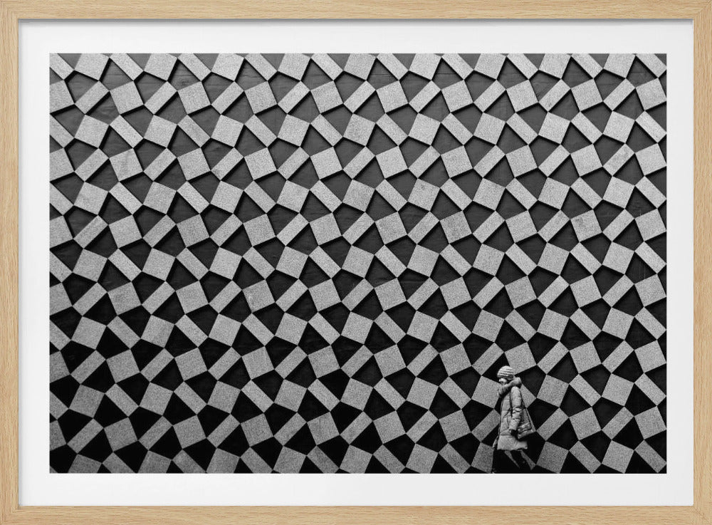 A black and white photograph of a lone person walking in front of a massive wall covered in a dizzying geometric pattern of tilted, textured gray quadrilaterals on a black background. The figure in the bottom right corner is dwarfed by the scale of the abstract architectural design. The image has a silver-gray frame. Artwork