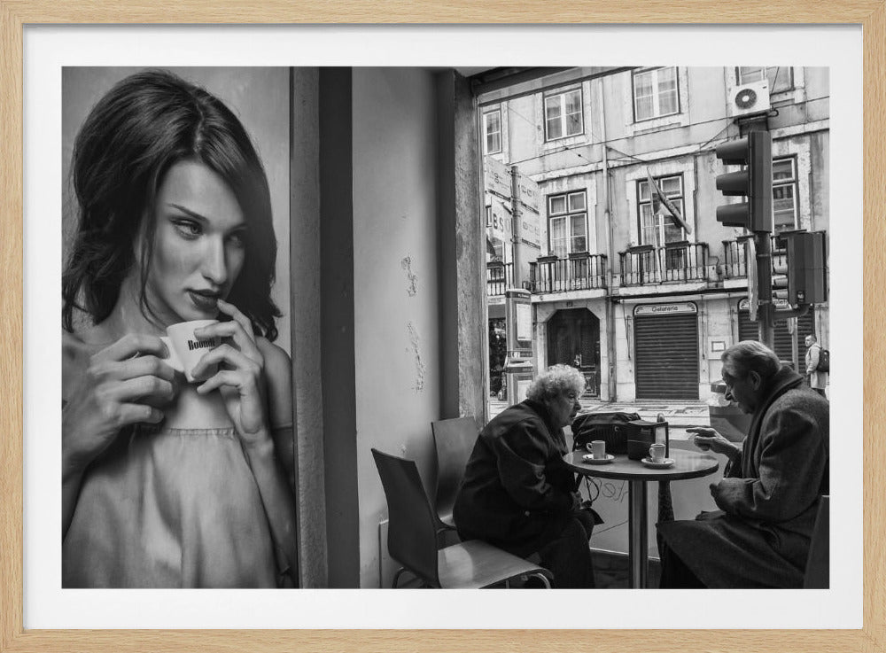 A framed, black and white photograph capturing a scene of juxtaposition. On the left, a large poster shows a glamorous young woman holding a coffee cup. On the right, seen through a cafe window, an elderly couple sits at a small table, also with coffee cups, engaged in conversation against a backdrop of an old European city street. Poster