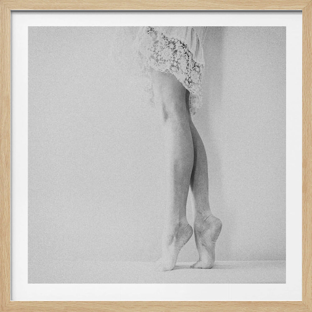 A framed, grainy black and white photograph of a dancer's legs and feet. The dancer stands on tiptoes against a white wall, wearing a delicate lace skirt. Poster