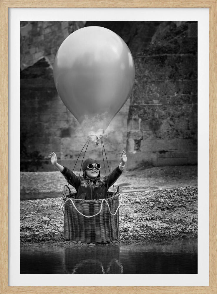 A whimsical black and white photograph of a young girl wearing an aviator cap and goggles, joyfully raising her arms in the air while standing in a wicker basket attached to a large balloon, mimicking a hot air balloon. The scene is set by the water with a stone wall in the background. Poster