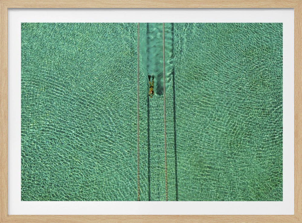 An aerial, top-down photograph of a lone swimmer in a lap lane of a large, turquoise-colored swimming pool. The water ripples from the sunlight, and the image is presented in a simple silver frame. Artwork