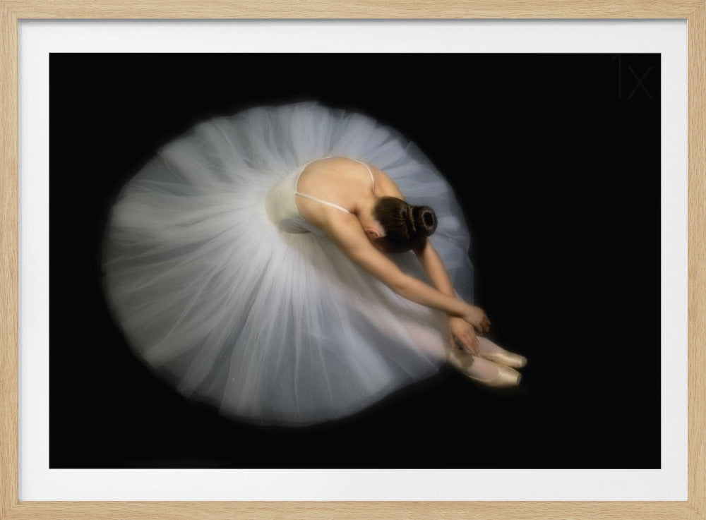 A high-angle photograph of a ballerina in a white tutu seated on a black background, bending forward in a graceful stretch. The tutu spreads out around her in a soft circle of light against the darkness. The image is presented within a silver frame. Print