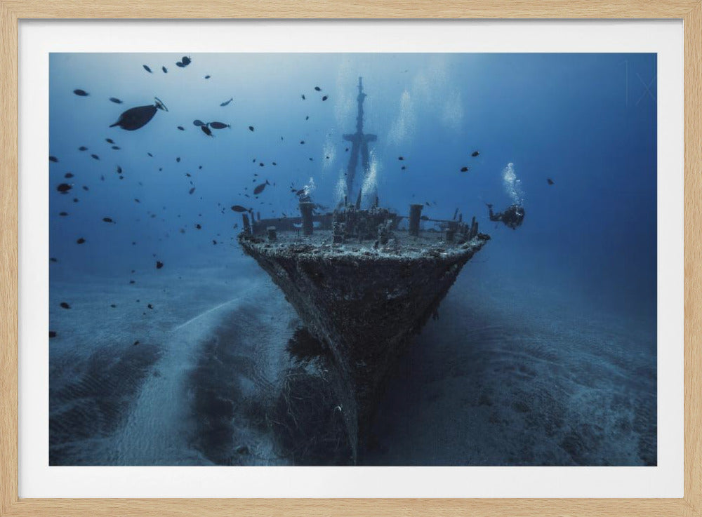 An underwater photograph of a large, sunken shipwreck resting on the sandy ocean floor. Two scuba divers are exploring the wreck, surrounded by schools of small fish in the deep blue water, with light filtering down from the surface. Print
