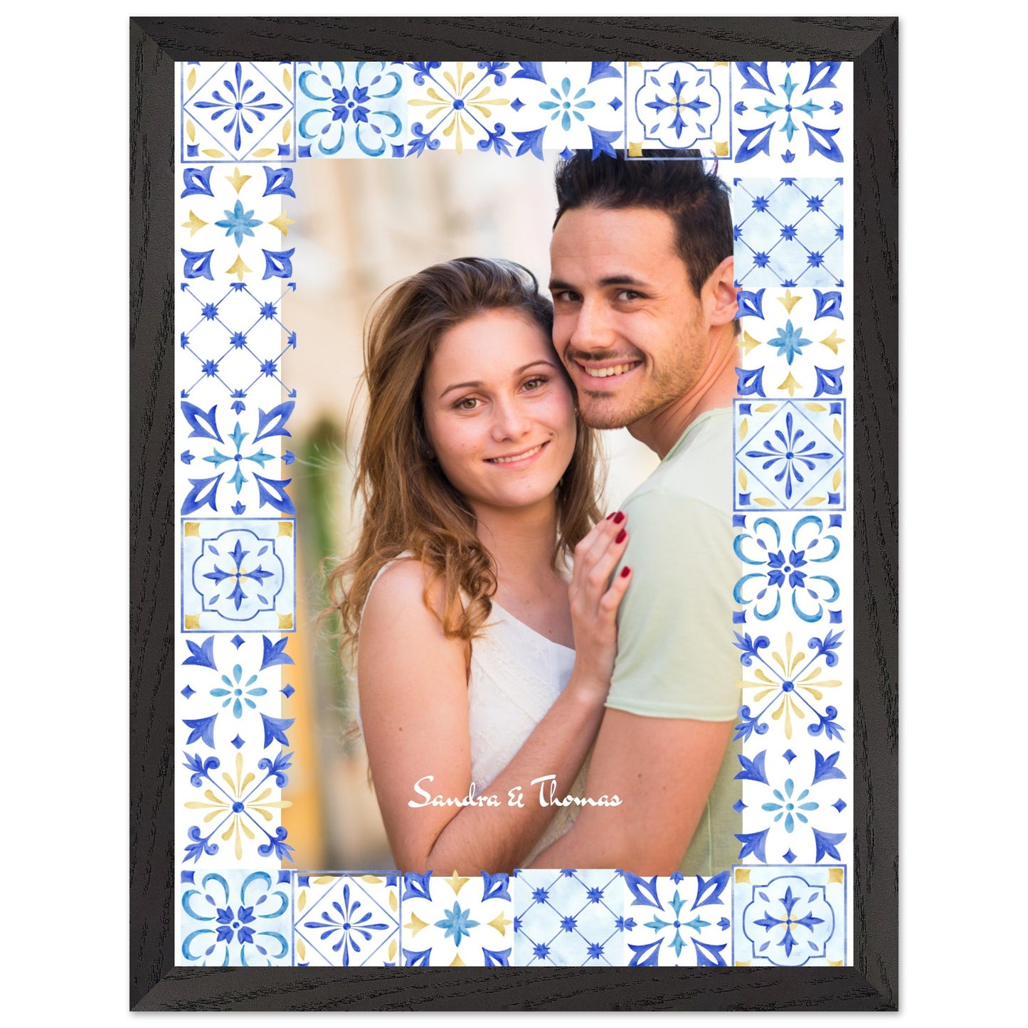 A personalized photo frame with a dark wood finish, featuring a photo of a smiling couple. The photo is bordered by a pattern of blue, yellow, and white Mediterranean-style tiles. The names 'Sandra & Thomas' are written on the photo in script. Print
