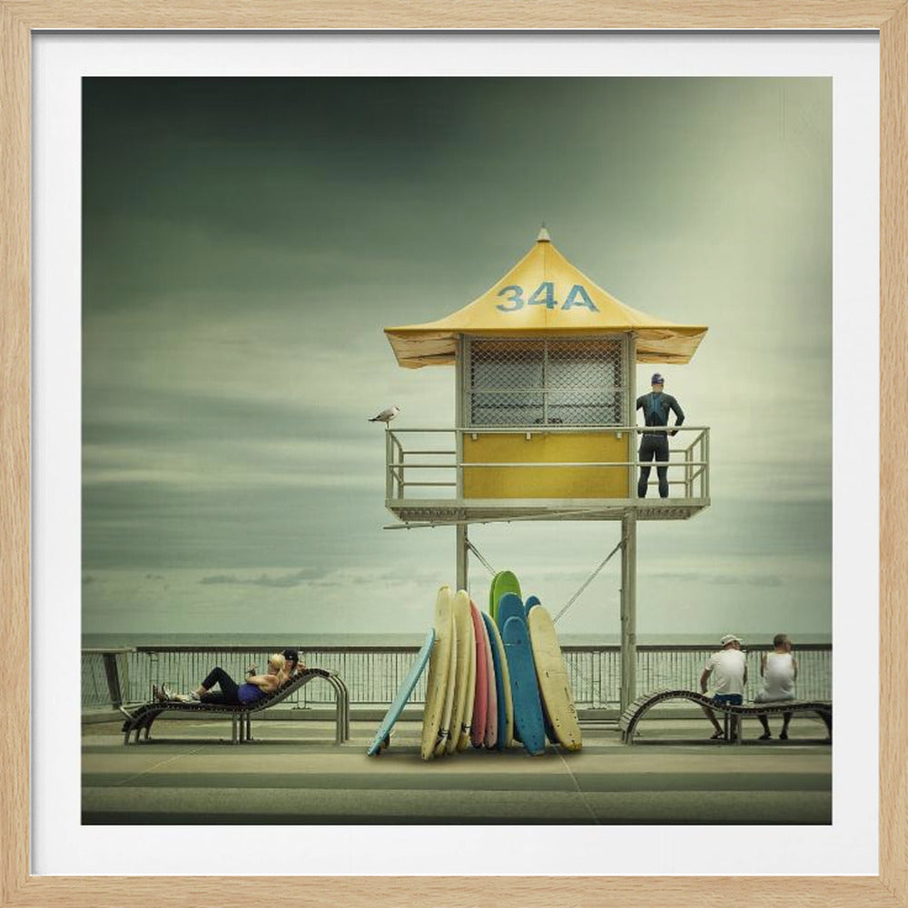 A vintage-style photograph of a yellow lifeguard tower labeled '34A' on a beach boardwalk under an overcast, greenish sky. A lifeguard in a wetsuit stands on the balcony, while a collection of colorful surfboards leans against the base. People are seen relaxing on benches nearby. Print