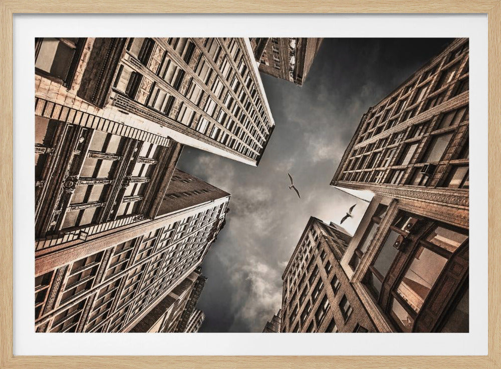 A dramatic, low-angle photograph looking up at the sky between several tall, sepia-toned skyscrapers that converge towards the center. Two birds are seen flying in the moody, dark grey sky. The image has a vintage feel and is presented in a silver frame. Wall Art