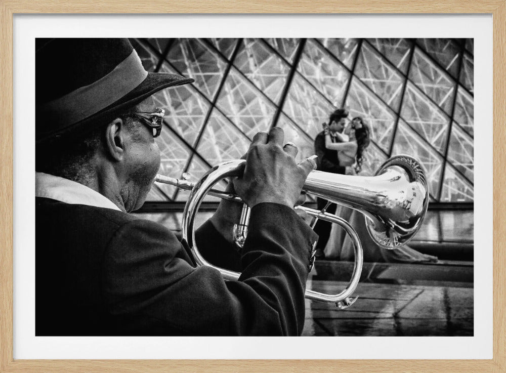 A black and white photograph from an over-the-shoulder perspective of an older Black man in a hat playing a silver flugelhorn. In the blurred background, a couple embraces in front of the glass panes of the Louvre Pyramid, creating a romantic Parisian scene. Print