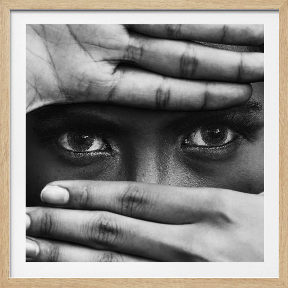A powerful black and white close-up photograph of a person's face, framed in light wood. Their hands cover most of their face, one over the forehead and the other over the mouth, revealing only their intense, striking eyes that gaze directly forward. Print