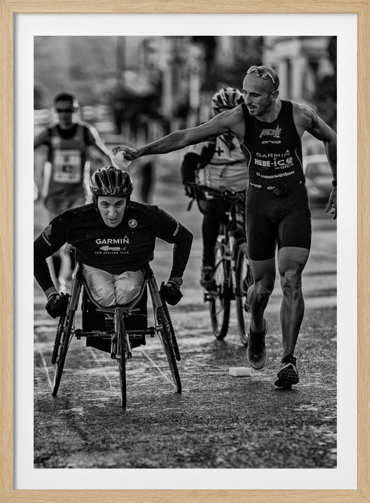 A dynamic black and white action photograph of a para-athlete in a racing wheelchair during a race. A running athlete beside him reaches out to hand him a sponge, showcasing a moment of camaraderie and support. Other competitors are blurred in the background on the wet asphalt. Print