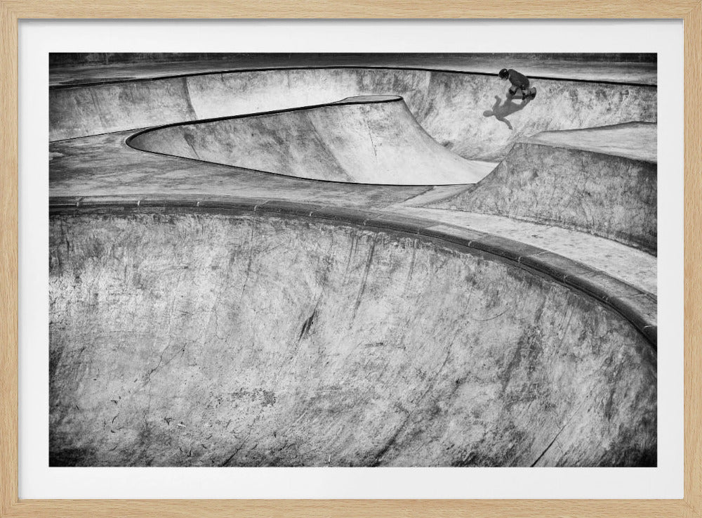 A high-angle, black and white photograph of a sprawling concrete skatepark with smooth, curving ramps. A single skateboarder is visible in the upper right, casting a distinct shadow on the ramp as they ride. The image is enclosed in a silver frame. Decor