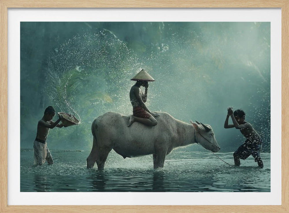 A serene photograph of three young boys playing in a river with a large water buffalo, one boy sits atop the animal while the others splash water, all set against a misty green backdrop and framed in silver. Print