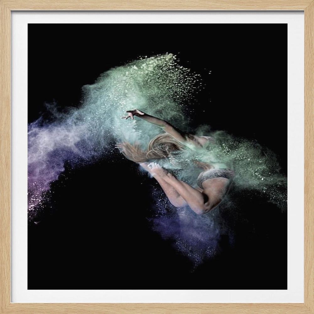 A female dancer is captured mid-air in an acrobatic leap against a solid black background. She is surrounded by a vibrant cloud of exploding green and purple powder, which traces the path of her movement and accentuates her athletic form. The image is presented within a light wood frame. Print