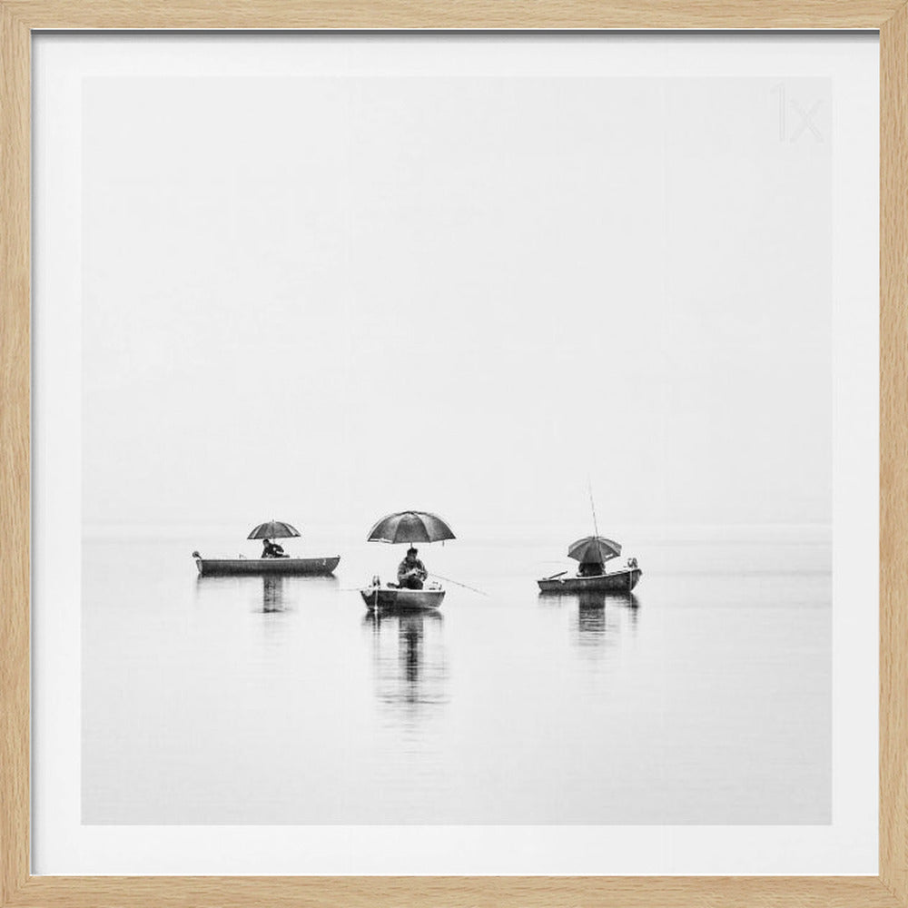 A minimalist black and white photograph of three fishermen in small boats on a calm, misty lake, each holding an umbrella. The image is framed in light wood. Artwork