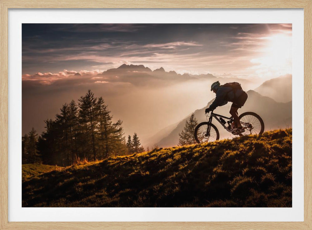 A mountain biker in silhouette rides down a grassy, sunlit hill, with a vast, misty mountain range in the background illuminated by a beautiful sunset. Wall Art