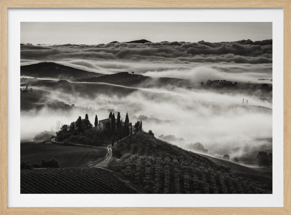 A framed, black and white photograph of a dramatic Tuscan landscape. A villa surrounded by cypress trees sits on a foreground hill, while thick fog blankets the rolling hills and valleys below. In the distance, a sea of clouds stretches towards a mountain range. Wall Art