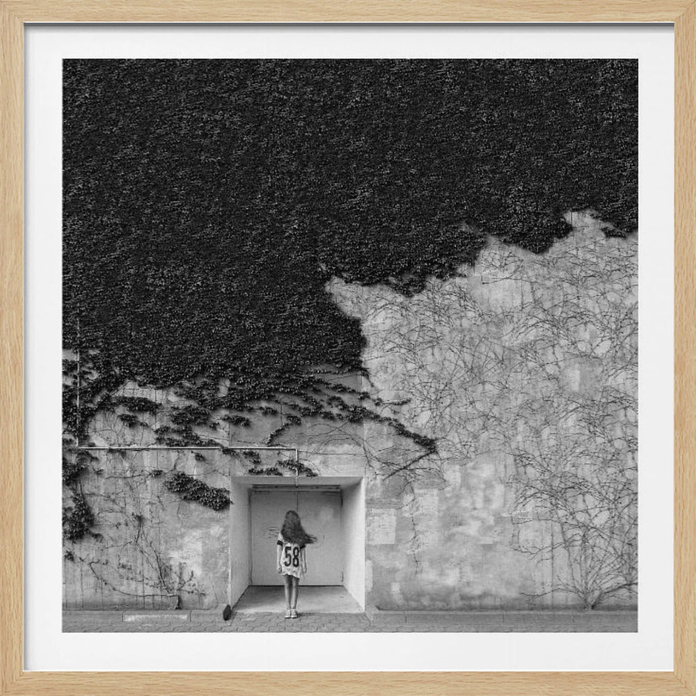 A framed, square black and white photograph showing a stark contrast on an ivy-covered wall. The top half is dense with dark leaves, while the bottom half is a lighter, textured wall. A young girl with long hair, seen from behind, stands in a recessed white doorway in the center. Wall Art