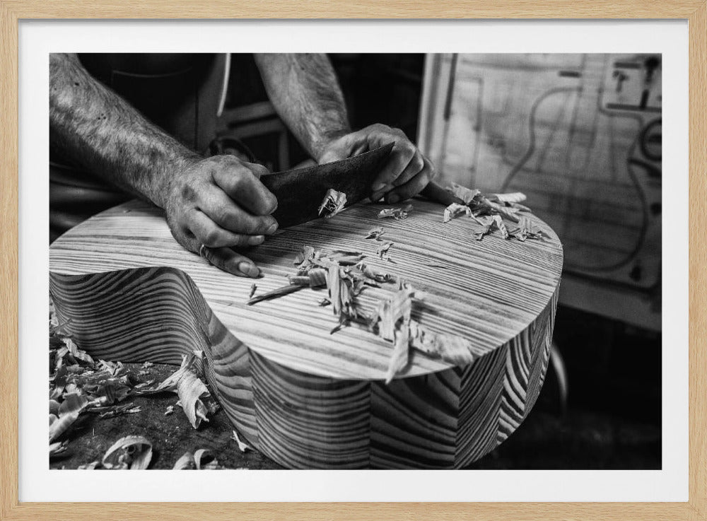 A black and white, framed photograph captures a close-up of a craftsman's hands using a tool to carve the body of a wooden guitar, with wood shavings littering the surface. Artwork
