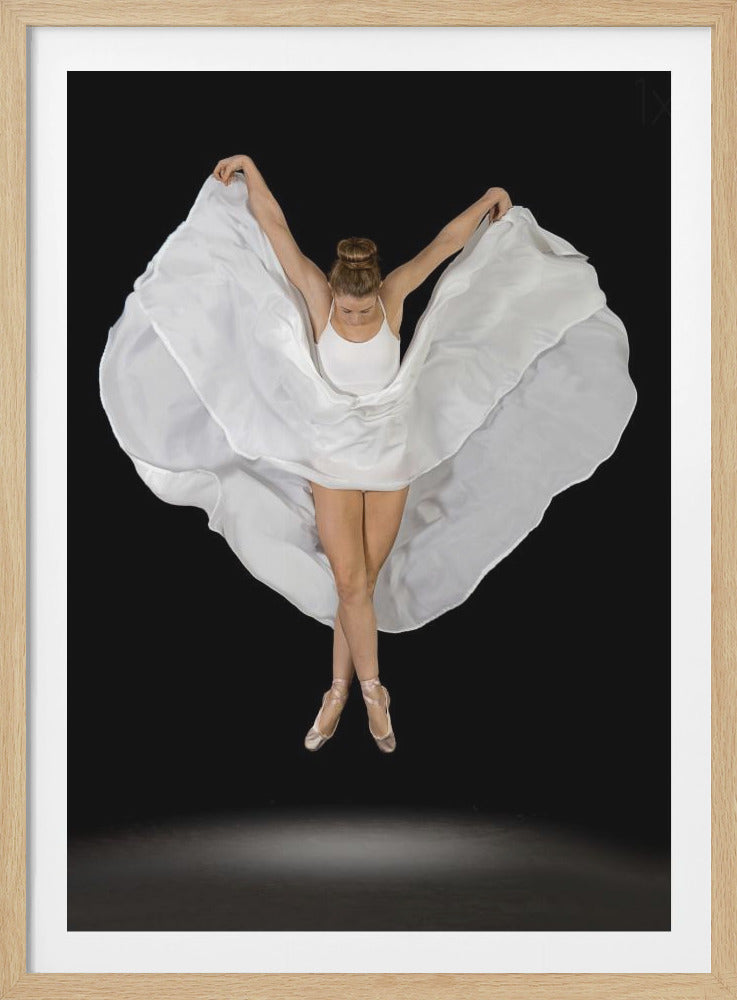A high-angle studio photograph of a graceful ballet dancer suspended mid-air against a solid black background. She wears a white leotard and pointe shoes, with her legs crossed. Her arms are outstretched, holding a large, flowing white fabric that creates a shape like wings around her. Decor