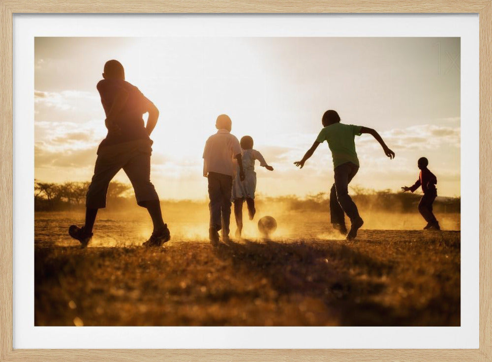 A low-angle, warmly lit photograph of five children silhouetted against a bright sunset as they play soccer on a dusty field, kicking up clouds of dust in the golden light. Decor