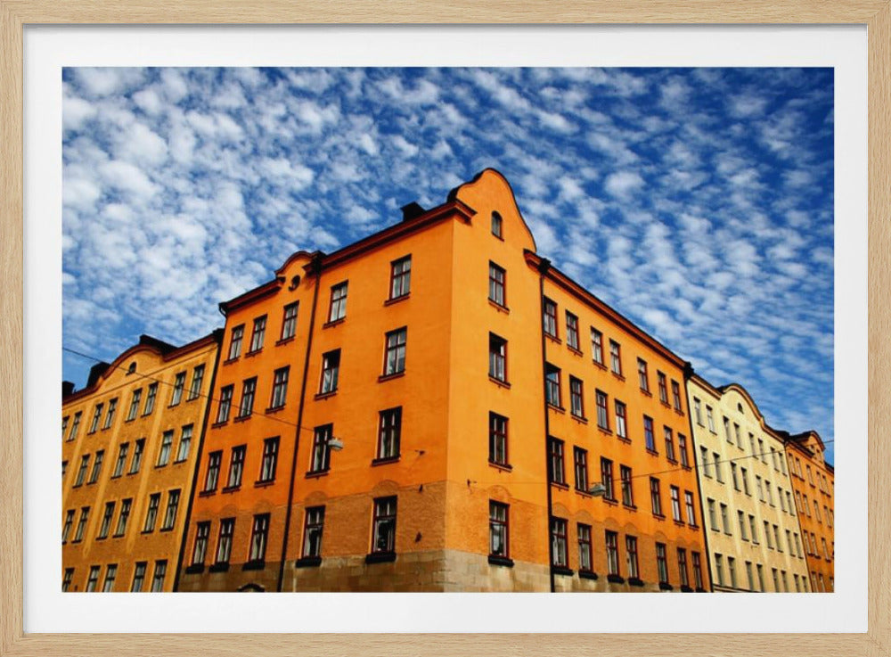 A low-angle shot of a vibrant orange apartment building against a dramatic blue sky filled with small, puffy white clouds. The building is on a corner, with another yellow-hued building visible next to it. The entire image is enclosed in a silver, brushed-metal style frame. Artwork