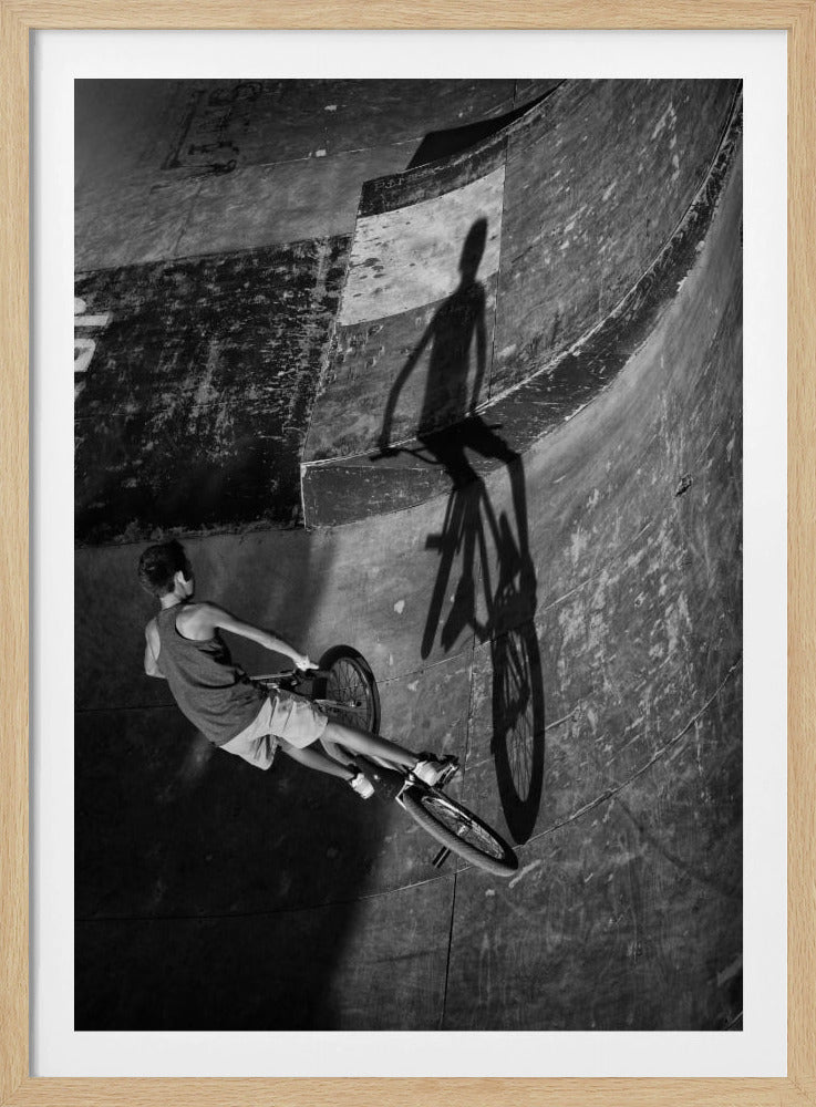 A dramatic black and white high-angle photograph of a BMX rider in a skate park. The rider is captured in motion on a curved ramp, while their long, stark shadow is cast on the worn, textured surface ahead, creating a powerful visual contrast. Poster