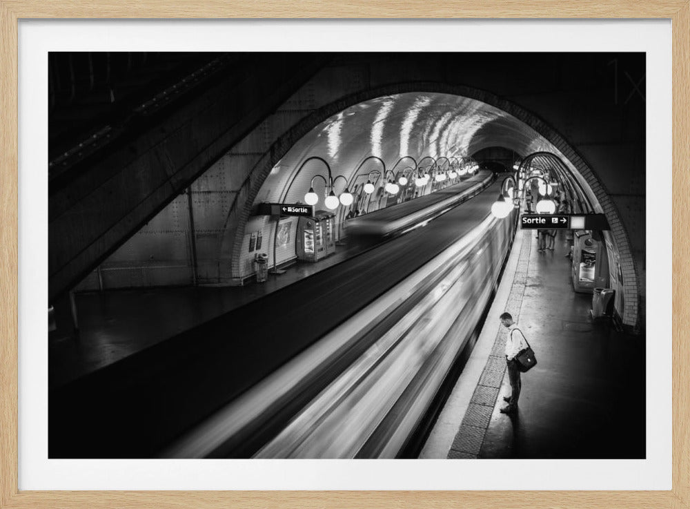 A black and white long-exposure photograph of a person waiting on a subway platform as a train speeds by, creating a motion blur. The photo is taken from a high angle in an arched tunnel station, with classic globe lights illuminating the scene. Poster