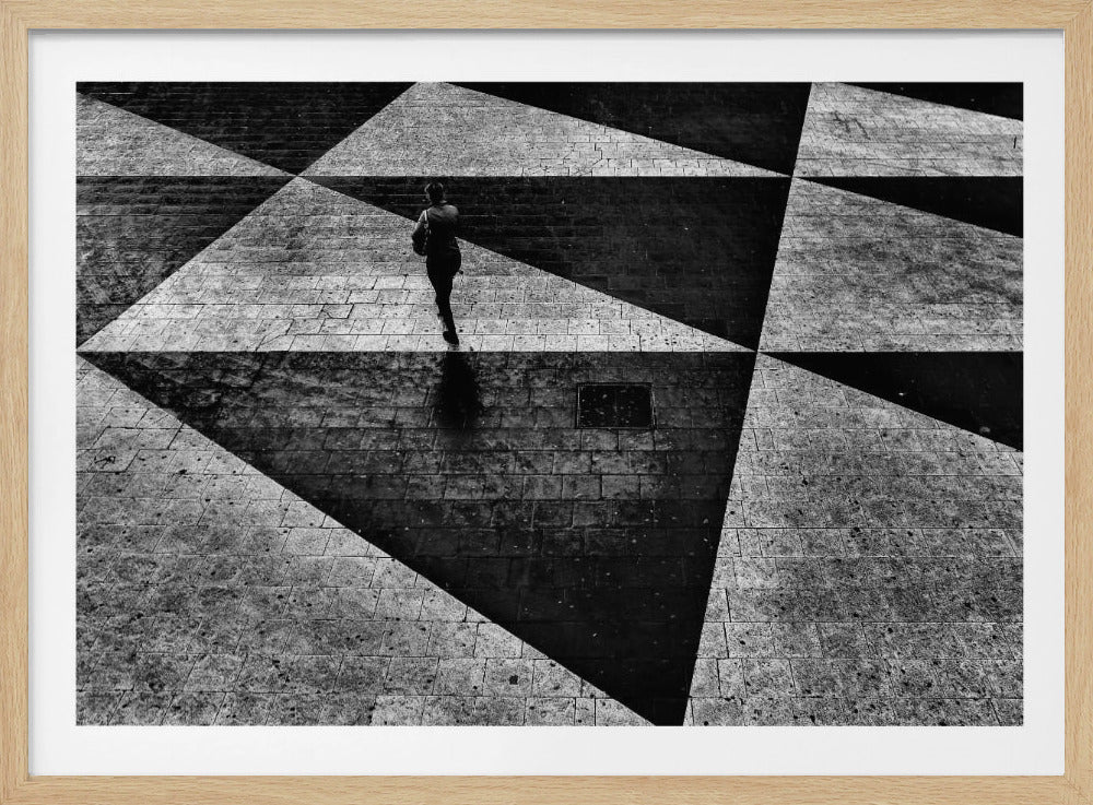 A dramatic, high-contrast black and white overhead photograph of a person walking across a paved ground with a striking geometric pattern of light and dark triangles created by strong shadows. The lone figure casts a long shadow, enhancing the minimalist and abstract composition, which is enclosed in a grey frame. Print