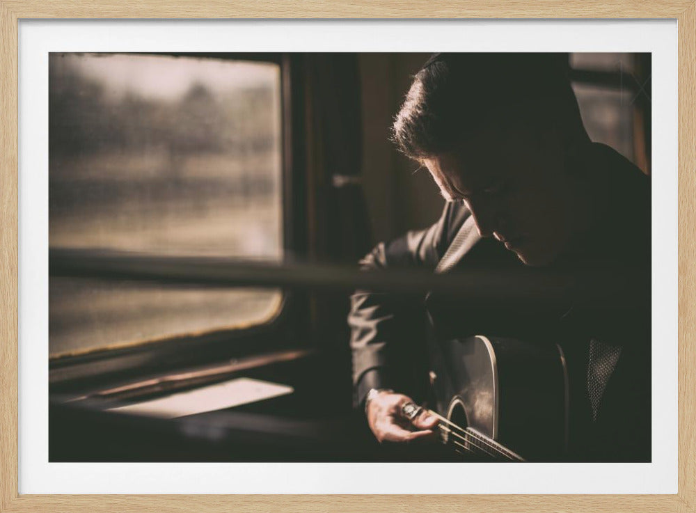 A moody, framed photograph showing a musician in profile, partially silhouetted against a window, deeply focused on playing an acoustic guitar. The scene is dimly lit, evoking a sense of introspection and travel. Artwork