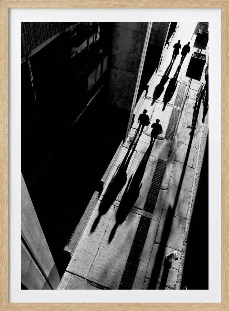 A dramatic high-angle, black and white photograph of several people walking on a sunlit sidewalk, casting long, stark shadows that stretch diagonally across the frame, contrasting with the deep black shadows of the adjacent building. Decor