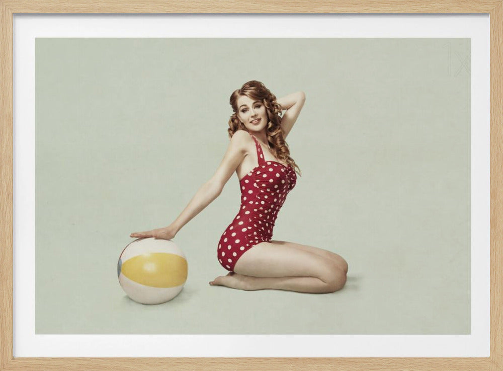 A framed vintage-style pin-up photograph of a woman with curly auburn hair, kneeling and posing in a red and white polka dot one-piece swimsuit. She smiles at the camera with one hand behind her head and the other resting on a yellow and white beach ball against a solid pale green background. Print