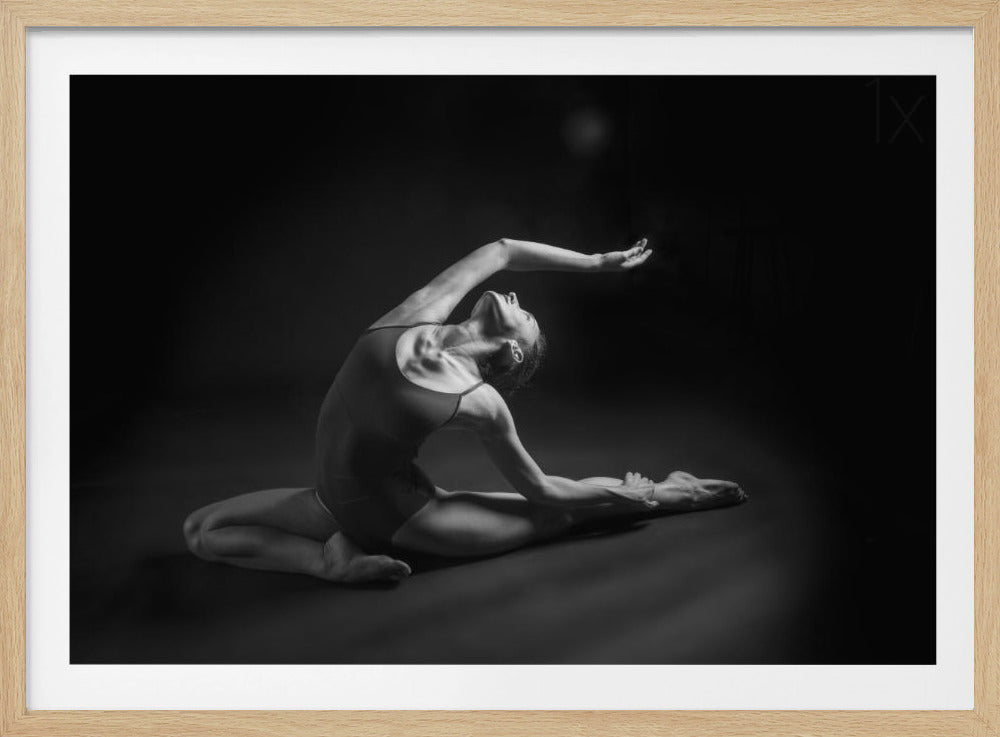 A dramatic black and white photograph of a female dancer in a leotard, performing a graceful stretch on a dark floor against a black background. She arches her back, looking up, with one arm extended beautifully above her head. Decor