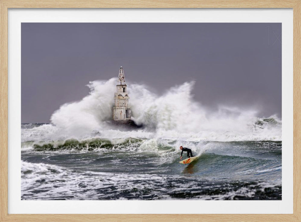 A framed photograph of a surfer in a black wetsuit riding a wave in a stormy sea. In the background, massive white waves crash against an old white lighthouse under a grey, overcast sky. Artwork