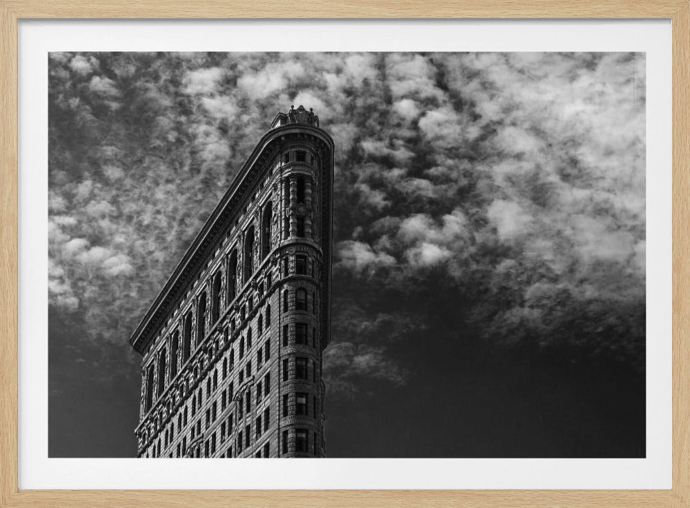 A dramatic low-angle black and white photograph of the Flatiron Building in New York City, with its sharp, triangular corner pointing towards a sky filled with fluffy, white clouds. The image is framed with a brushed silver border. Print