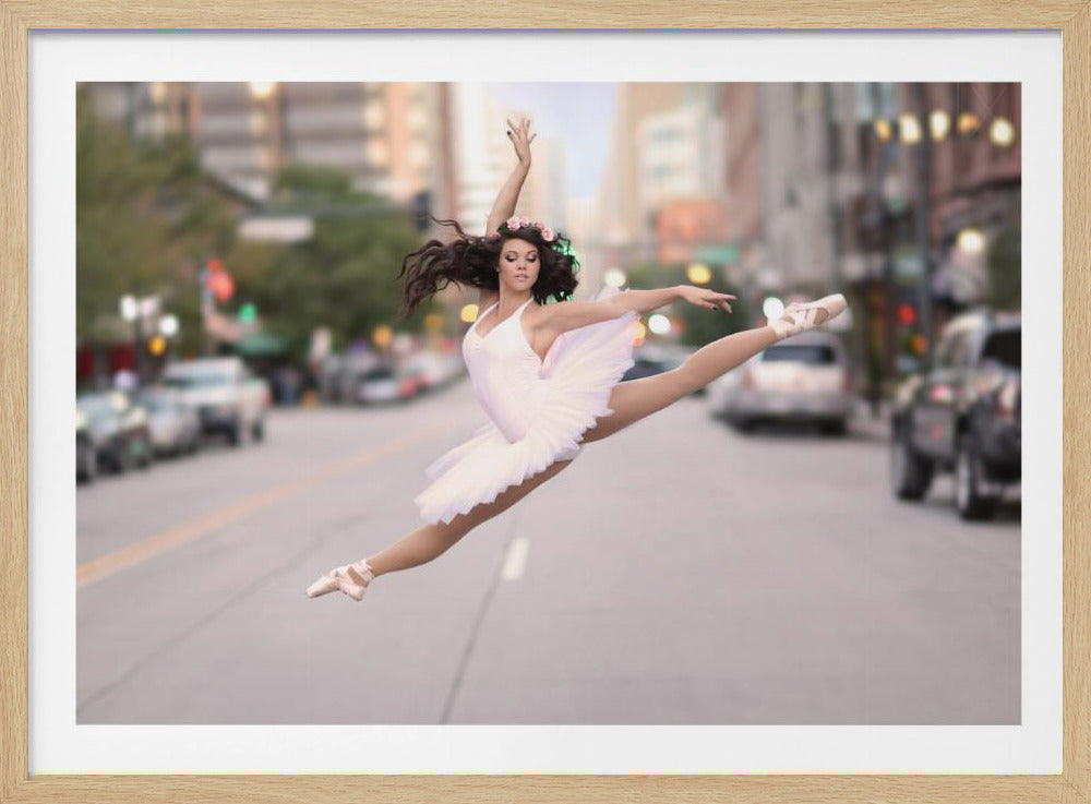 A ballerina in a white tutu and pointe shoes leaps into a split mid-air in the middle of a blurred city street, her arms extended gracefully. The image is set within a silver frame. Print
