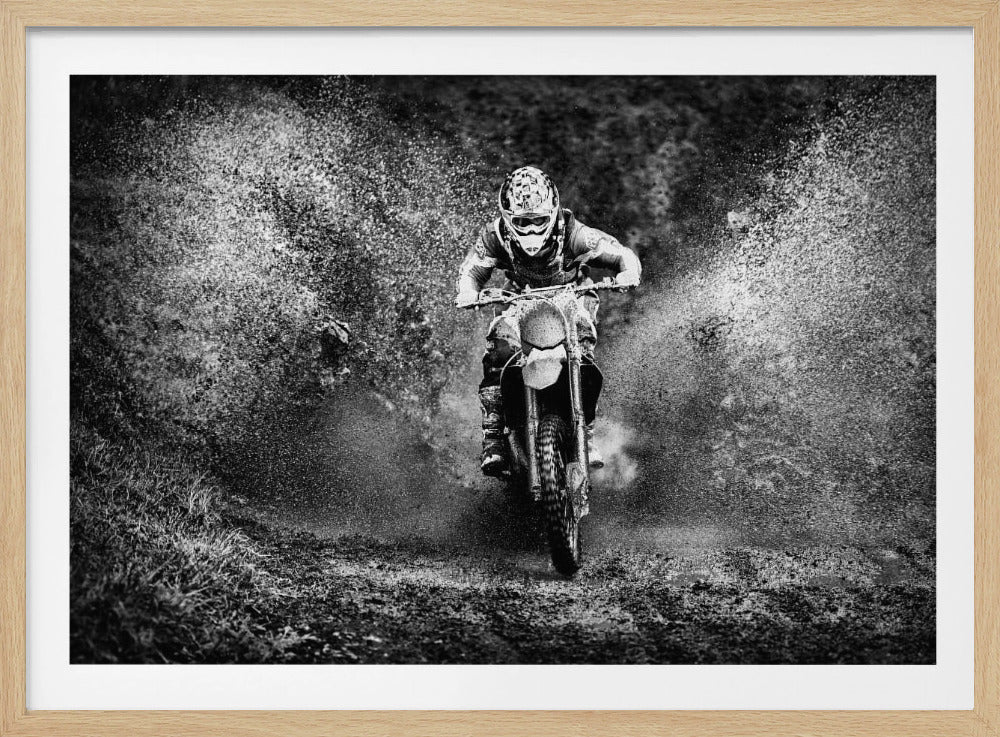 A dramatic black and white action shot of a motocross rider in full gear, riding head-on through a muddy puddle, creating a massive splash of water and dirt on both sides. The image is captured in a grainy, high-contrast style and is enclosed in a silver frame. Print