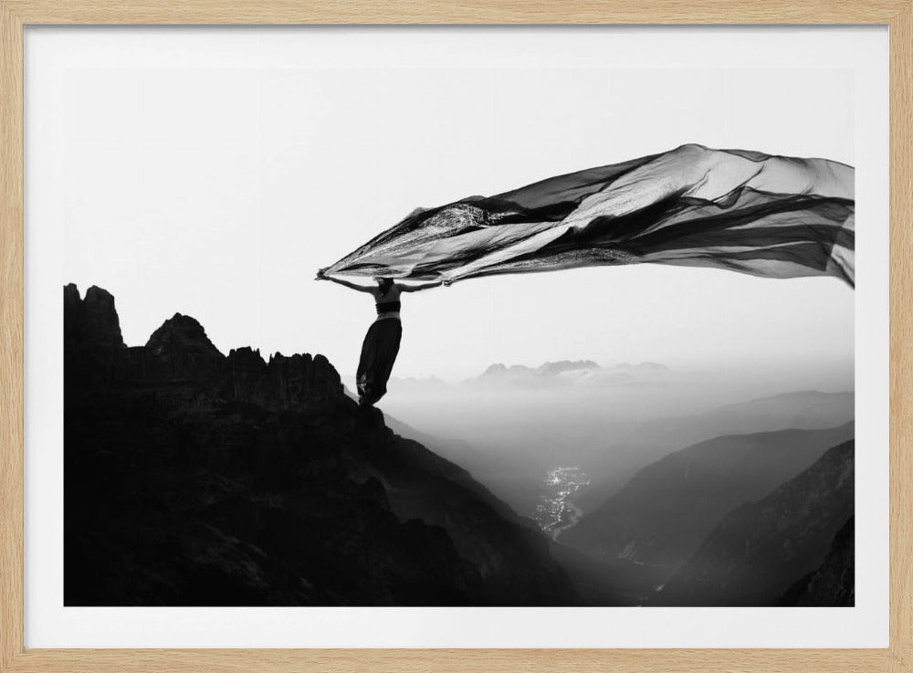 A dramatic black and white photograph of a woman standing on the edge of a mountain peak. She holds up a massive piece of dark, sheer fabric that billows in the wind, stretching across the sky. Below her, a vast, misty mountain range and a deep valley with city lights are visible. Wall Art