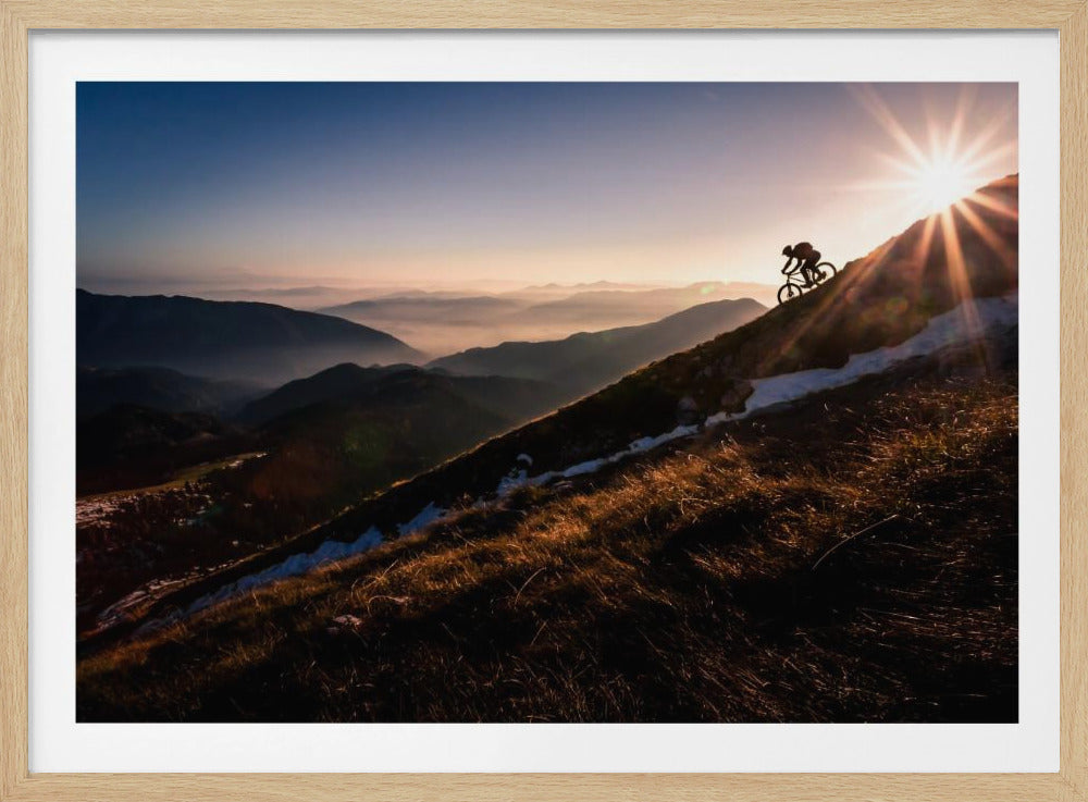 A silhouette of a mountain biker pushing their bike up a steep mountainside against a brilliant sunrise. The sun is cresting the peak, creating a starburst effect with long light rays. In the background, layers of mountains and valleys are filled with a soft morning mist under a clear gradient sky. Artwork