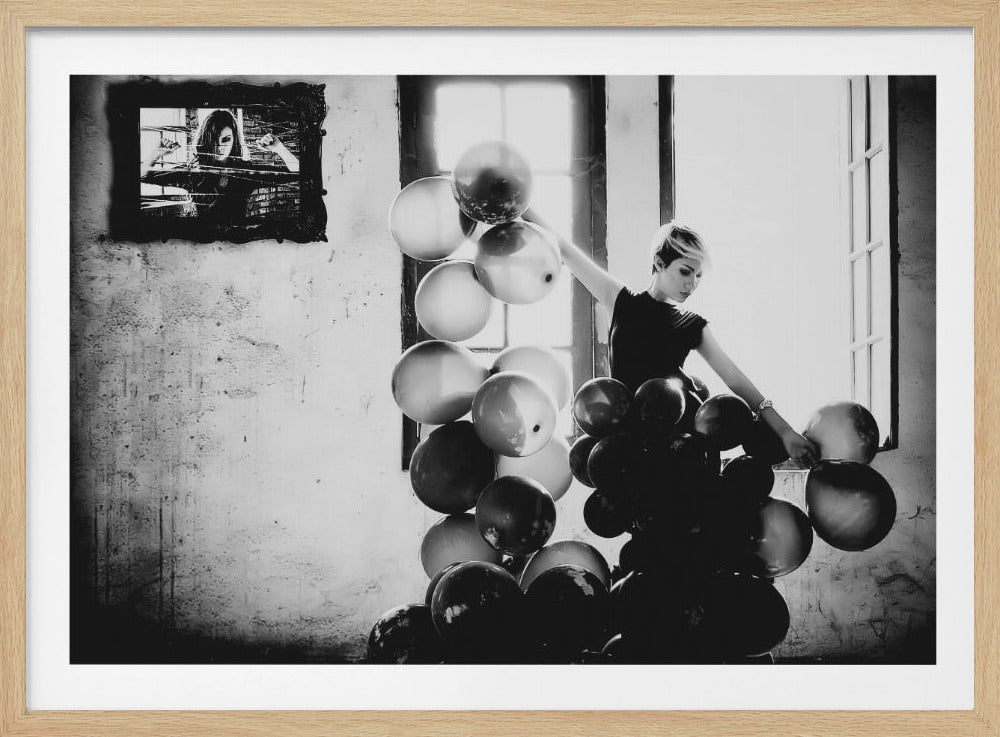 A framed, black and white artistic photograph of a woman with short blonde hair in a dark dress, standing by a bright window. She is surrounded by a large cluster of black and white balloons. On the distressed wall to the left, there is a dark, ornate frame holding a picture of another woman looking intensely forward. Wall Art