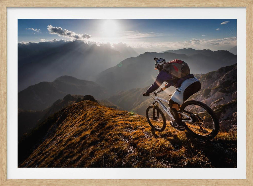 A mountain biker navigates a narrow, golden-lit ridge against a backdrop of a vast mountain range. The sun is breaking through clouds in the distance, casting dramatic rays of light across the epic landscape. Print