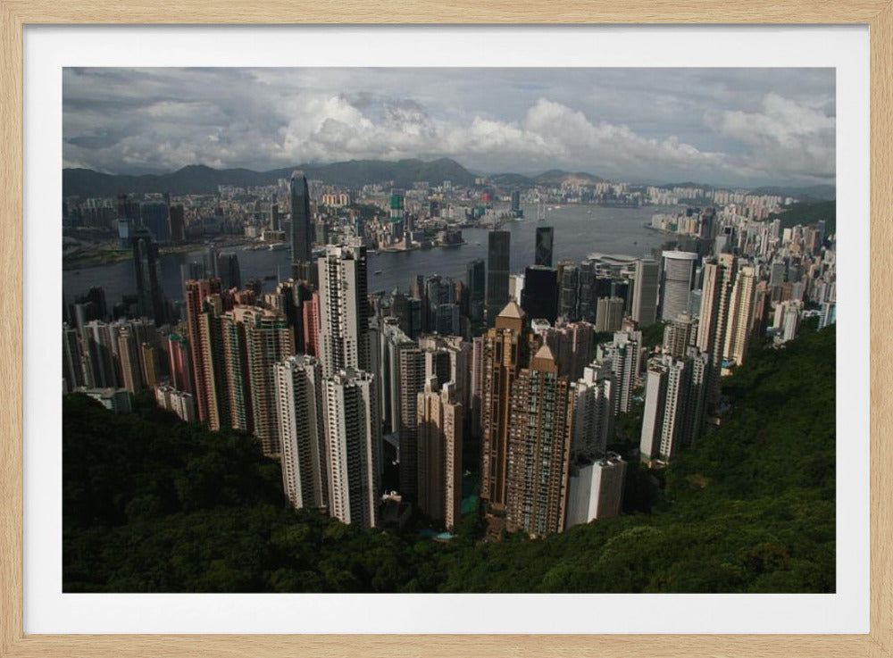 A high-angle photograph of the Hong Kong cityscape, viewed from a lush green peak. Numerous skyscrapers fill the frame, overlooking a bustling harbor with mountains in the distant background under a cloudy sky. The entire image is presented within a silver, brushed-metal frame. Artwork